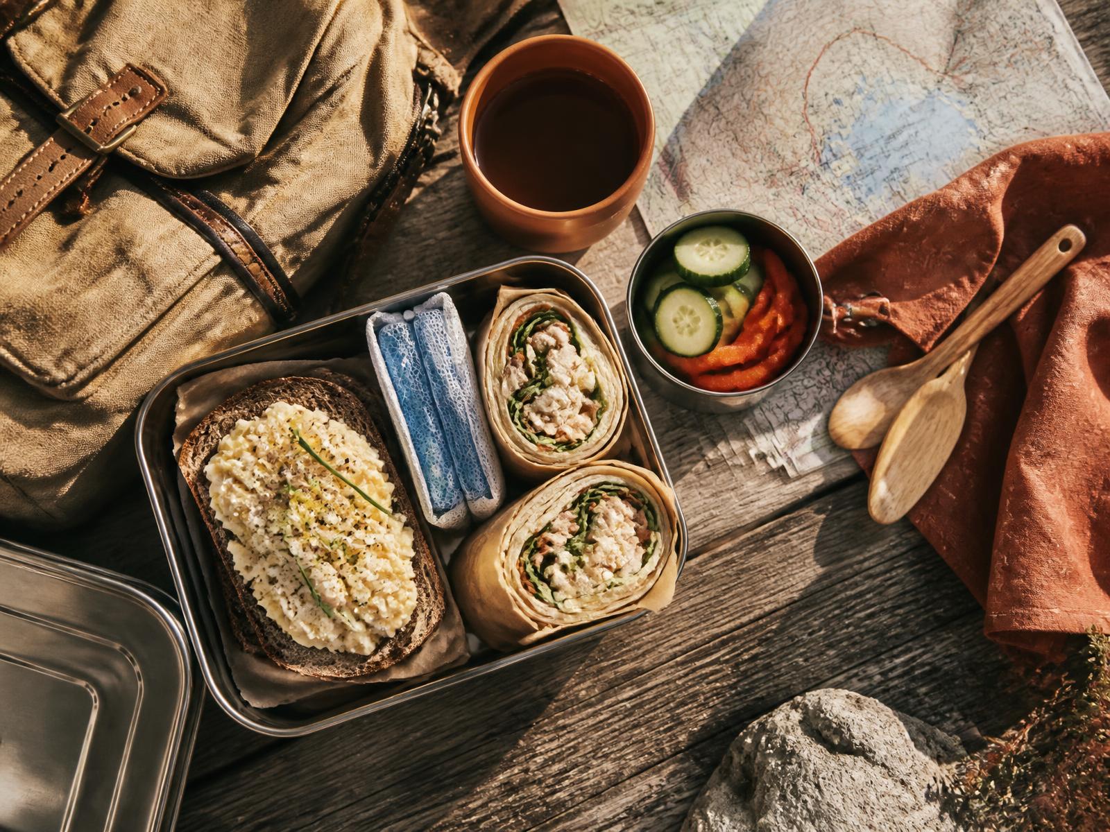 A packed travel meal of egg salad, wraps and vegetables in a stainless tin beside a backpack and a paper map